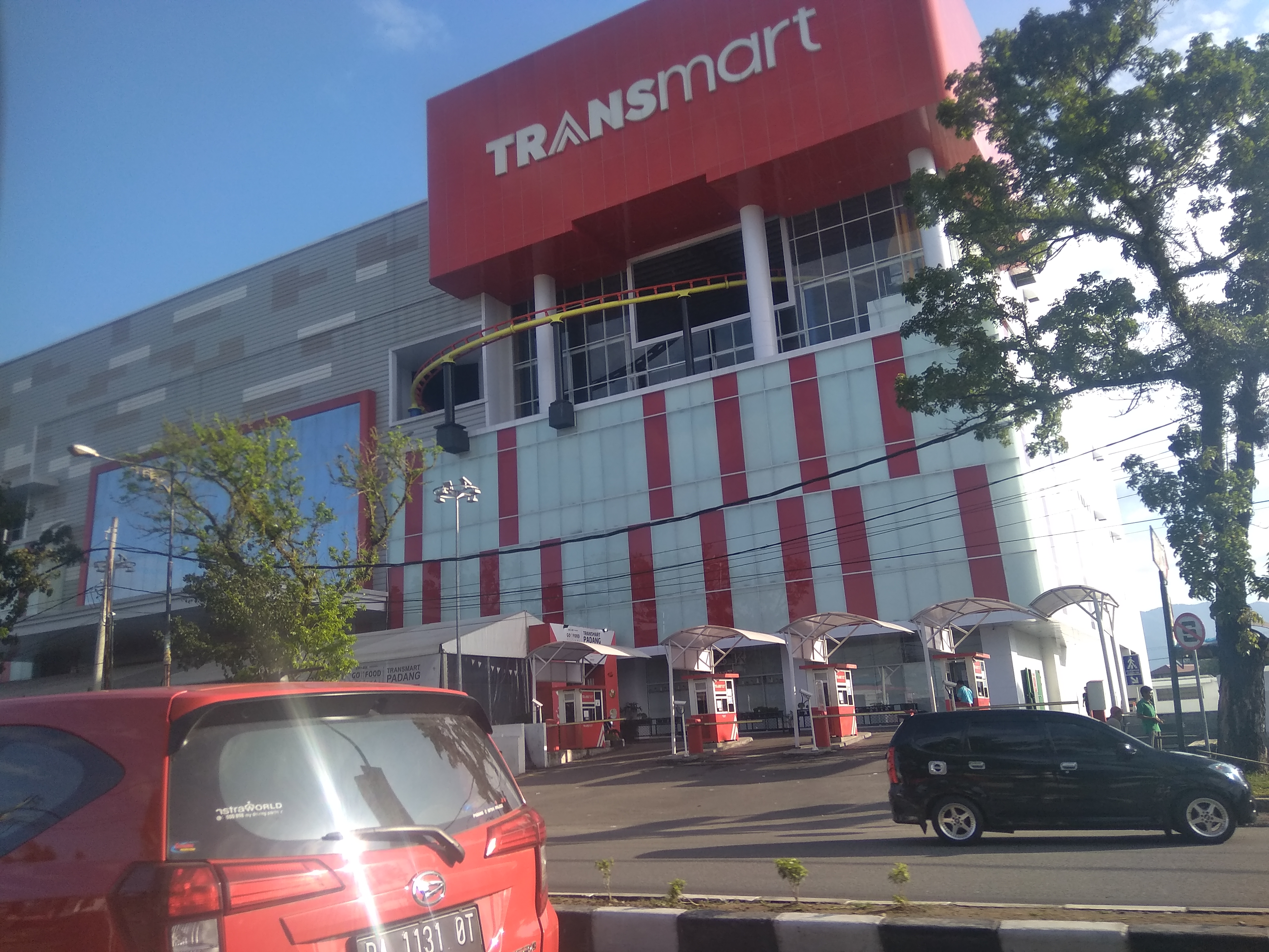 Gedung Transmart dengan logo merah di bagian atas, memiliki fasad putih dan merah, dengan beberapa mobil parkir di depannya dan pohon di sekitarnya.